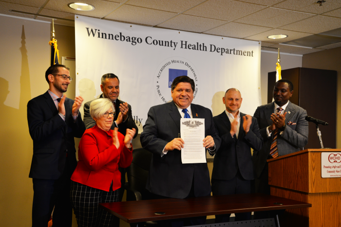 Picture of Governor Prizker holding up signed Executive order with five supporters clapping